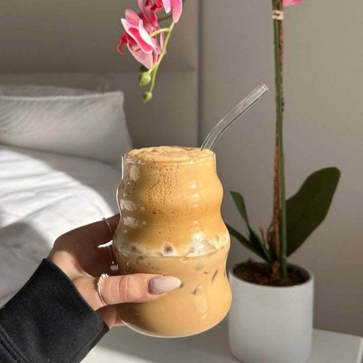 Hand holding a curved glass of iced coffee with a straw, surrounded by a plant and pink flowers.